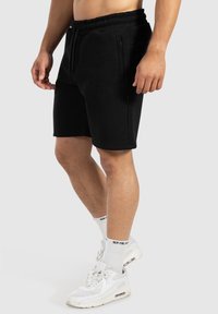 Pantalones cortos negros, hasta la rodilla, hechos de una tela suave, con cinturilla elástica, bolsillos con cremallera en los laterales y una textura lisa. Combinados con zapatillas blancas.