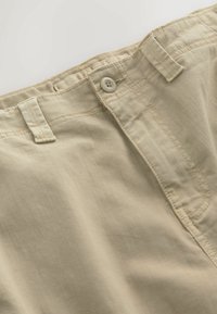 Pantalon en coton beige au niveau de la taille montrant un bouton, des passants pour ceinture et des coutures détaillées sur fond uni.