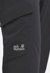 Primo piano di pantaloni da esterno scuri con una tasca a pattina e il logo Jack Wolfskin con impronta di zampa sulla coscia.