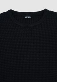 Maglione nero lavorato a maglia con superficie strutturata e scollo rotondo. Etichetta visibile con informazioni su marca e taglia.