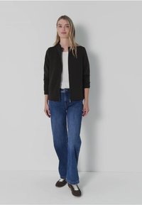 Czarna kurtka zapinana na zamek na białej koszuli, w połączeniu z niebieskimi dżinsami w stylu dzwonów i brązowymi trampkami. Gładkie faktury i swobodny design.