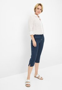 Camicia bianca in lino a maniche lunghe, leggera, abbinata a pantaloni blu navy a lunghezza ginocchio con coulisse, dotati di aperture laterali e dettagli dorati, indossata con sandali bianchi.