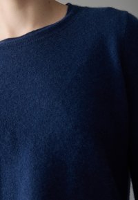 Gros plan sur un torse portant un pull en tricot bleu foncé avec un col rond, sur un fond neutre.