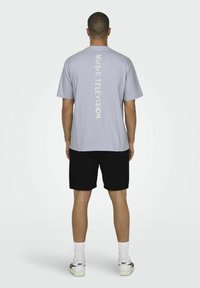 Lyseblå t-shirt med "MUSIC TELEVISION" trykt vertikalt i hvidt på ryggen, matchet med sorte shorts og hvide sneakers.