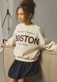 Cremefärgad sweatshirt med "BOSTON" tryckt i mörkröd, i kombination med en marinblå kjol. Tröjan har ribbade ärmslut och en avslappnad passform, vilket framhäver enkla linjer.