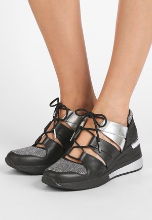 Pieds chaussés de baskets compensées noires et argentées à lacets avec tissu en maille et sangles métalliques à la cheville sur fond blanc.
