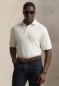 Hombre con gafas de sol oscuras, camisa polo blanca de manga corta, vaqueros oscuros con cinturón marrón y reloj con correa de cuero marrón.