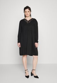 Vestido preto de manga longa com decote em V e detalhes em renda, apresentando uma cintura franzida e um corte relaxado, combinado com sapatos de salto alto pretos.