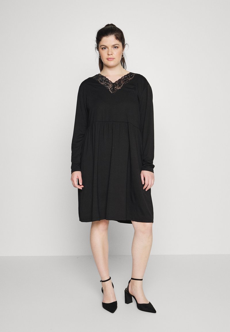 Vestido preto de manga longa com decote em V e detalhes em renda, apresentando uma cintura franzida e um corte relaxado, combinado com sapatos de salto alto pretos.