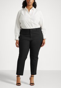 Camicia bianca a maniche lunghe, abbinata a pantaloni neri su misura. Il modello indossa sandali neri con cinturino, creando un look professionale.