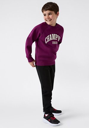 Sudadera morada con estampado "CHAMPION 1919", combinada con pantalones negros y zapatos negros con suelas blancas y acentos rojos. Textura de tela suave.