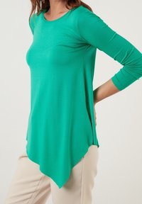 Grön långärmad topp med rundad halsringning och asymmetrisk nederkant. Tillverkad av mjukt, stretchigt tyg, med en figursydd överdel och l ös drapering.