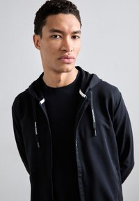 Felpa nera con zip e cappuccio regolabile, caratterizzata da dettagli bianchi e il logo "Armani Exchange". Indossata sopra una t-shirt nera.