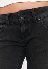 Jeans in denim nero con un design aderente, due bottoni frontali e tessuto testurizzato. Gli accessori includono un piccolo dettaglio metallico in vita.