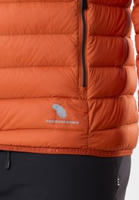 Oranje donsjack met een gequiltde textuur, voorzien van een ritssluiting, logo-details en elastische afwerking aan de onderkant, gecombineerd met zwarte broeken.