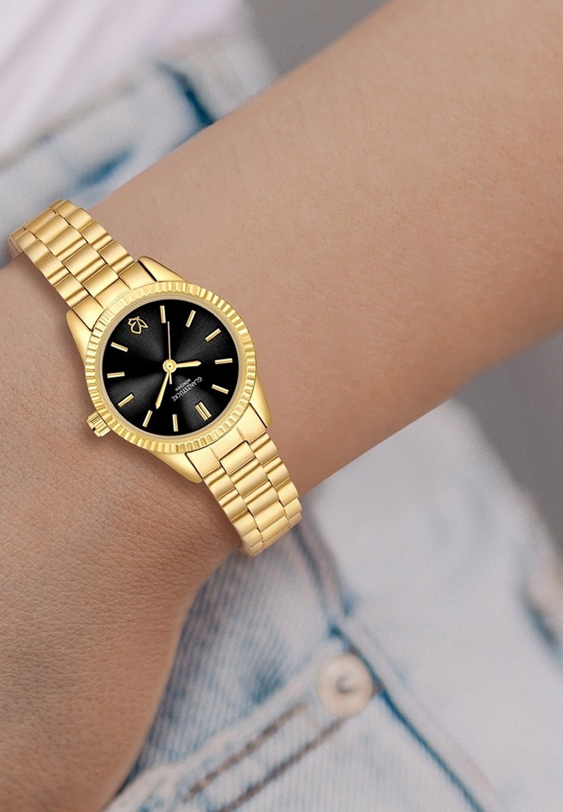 Zelts rokas pulkstenis ar apaļu melnu ciparnīcu, zelta stundu rādītājiem un teksturētu rāmi, kas apvienots ar zelta metāla saites sprādzi.