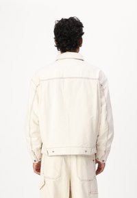 Chaqueta de mezclilla blanca con costuras visibles y botones a presión. Diseño casual con corte recto y cuello clásico.