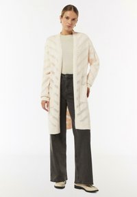 Langer, offener Cardigan in Creme mit einem Zickzack-Open-Knit-Muster, kombiniert mit einem hellen Oberteil und dunklen, weit geschnittenen Jeans. Mit seitlichen Taschen akzentuiert.