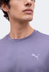 Νέος άνδρας με λιλά μπλούζα Puma, ορατός από τον λαιμό μέχρι το στήθος, κοιτάζει ελαφρώς προς τα πάνω και στο πλάι.