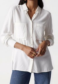 Chemise blanche légère à boutons avec tissu texturé, deux poches poitrine et col. Portée avec un jean denim bleu.