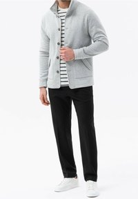 Chaqueta de punto gris con cuello alto, cierre de botones, dos bolsillos laterales; llevada sobre una camisa a rayas, combinada con pantalones negros y zapatos blancos.