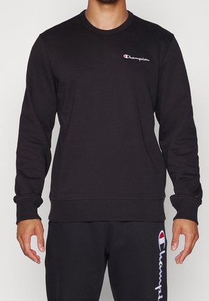 Homme portant un sweat-shirt noir Champion et un pantalon noir assorti avec des logos Champion visibles sur la poitrine et la jambe.