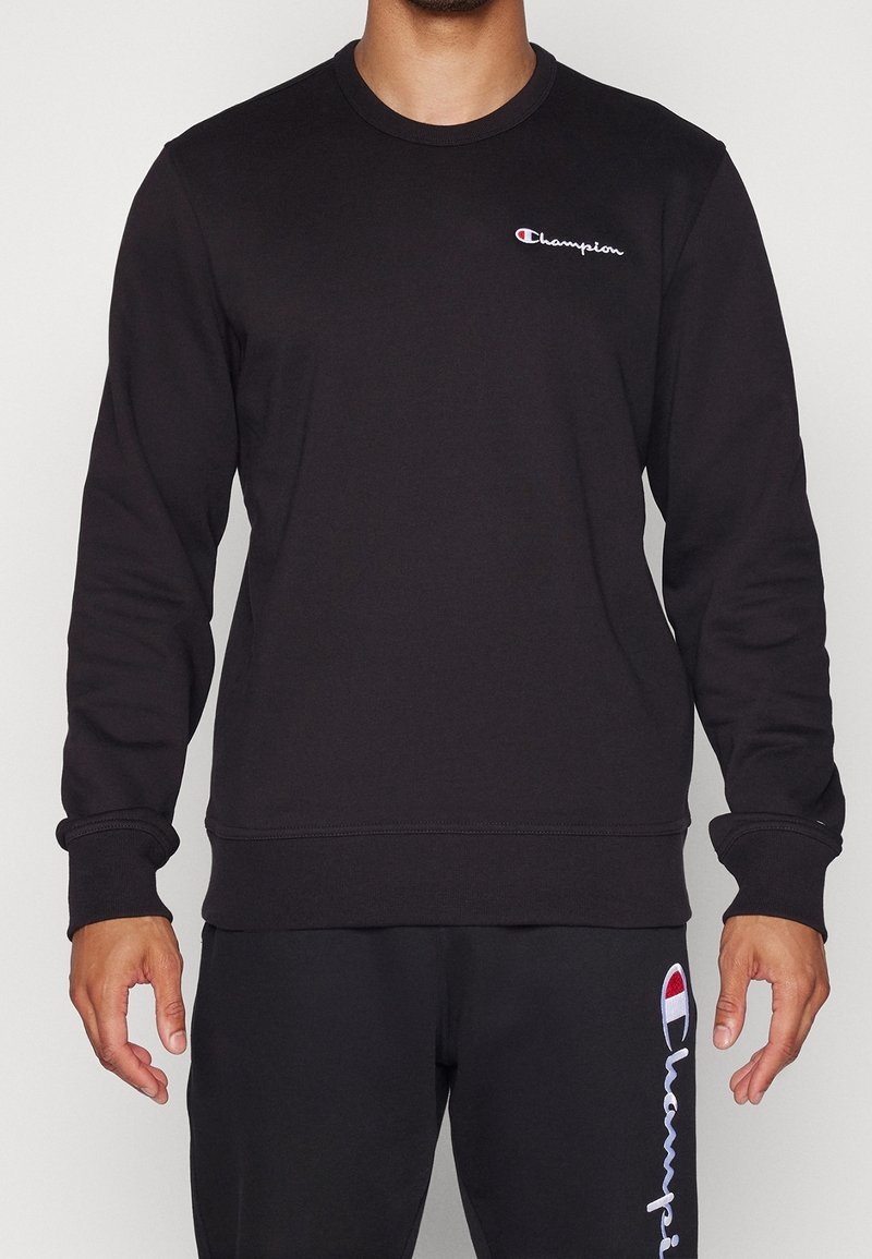 Homme portant un sweat-shirt noir Champion et un pantalon noir assorti avec des logos Champion visibles sur la poitrine et la jambe.