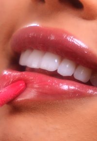 Close-up van lippen waarop een glanzend, dieproze lipproduct wordt aangebracht. Witte, rechte tanden zijn zichtbaar, wat de uitstraling versterkt.