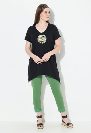 Túnica negra con escote en V y bolsillo redondo decorado, combinada con leggings verdes con detalle de encaje en el dobladillo y sandalias negras de cuña.