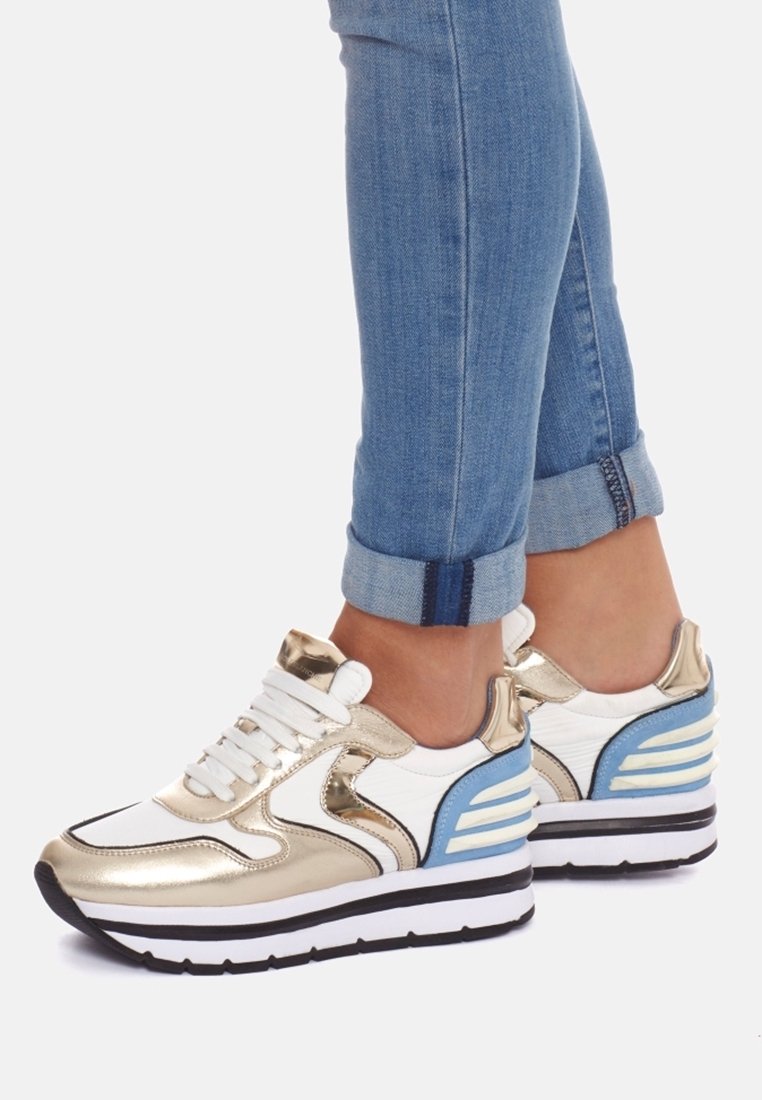 Sneaker metallici oro e bianchi con accenti blu, dotati di una suola spessa e un design testurizzato, abbinati a jeans arrotolati.