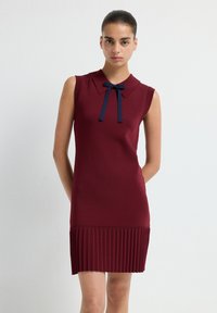 Abito senza maniche color bordeaux con colletto e dettaglio a fiocco blu navy; la texture plissettata all'orlo aggiunge interesse visivo. Finitura in tessuto liscio.