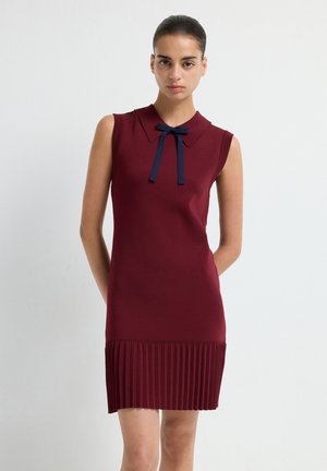 Robe sans manches bordeaux avec un col et un détail de nœud bleu marine ; la texture plissée à l'ourlet ajoute un intérêt visuel. Finition en tissu lisse.