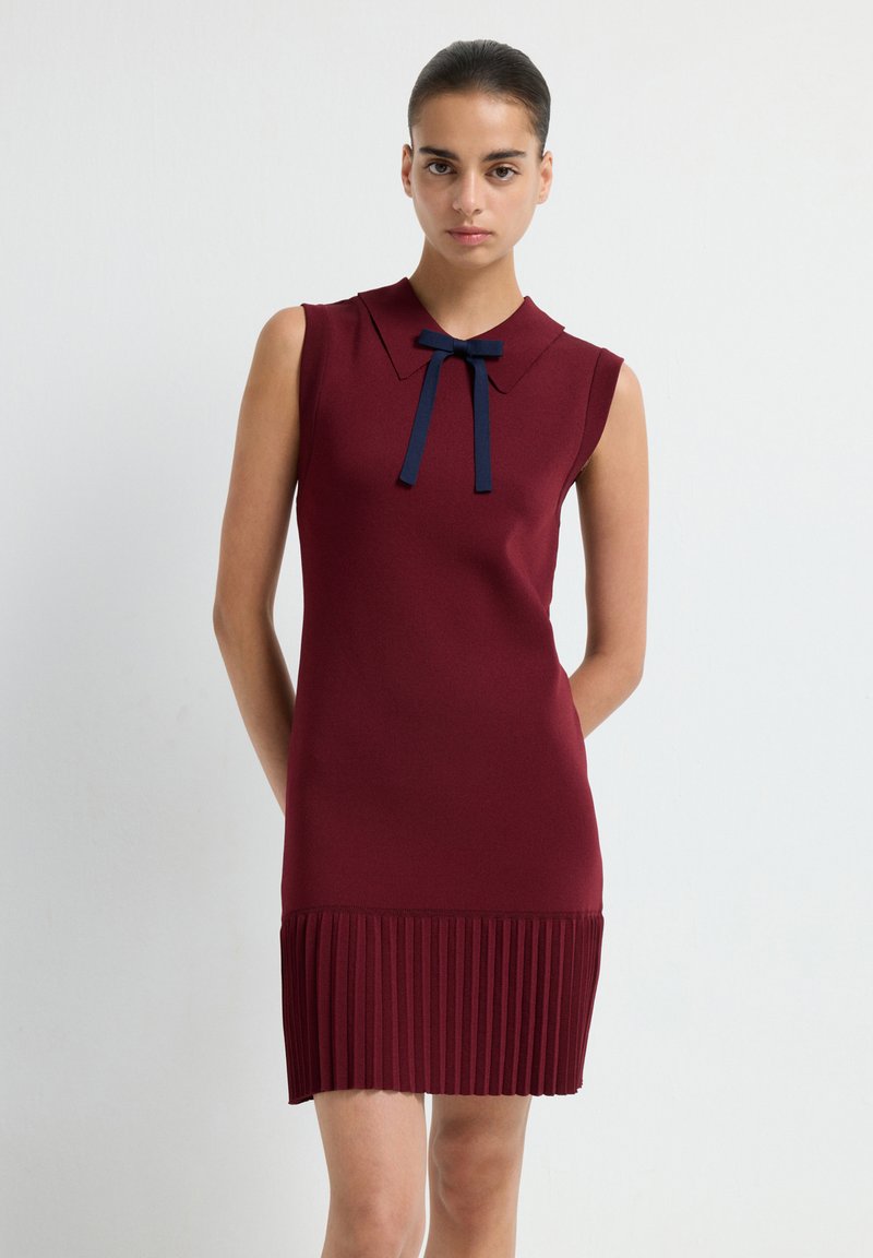 Abito senza maniche color bordeaux con colletto e dettaglio a fiocco blu navy; la texture plissettata all'orlo aggiunge interesse visivo. Finitura in tessuto liscio.