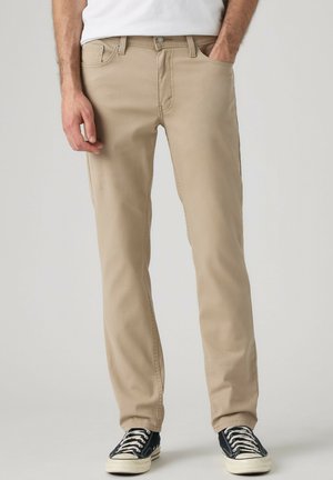 Mand iført beige bukser med hænderne i lommerne og sorte og hvide sneakers, der står mod en ensfarvet baggrund.