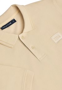 Beige polo skjorta med texturerad tyg, tre-knapps knäppning och en broderad "POLO CLUB" logotyp på bröstet. Kortärmad och med ribbad krage.