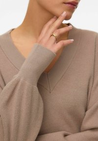 Pull en maille beige avec un col en V et de longues manches côtelées ; la main du modèle repose sur son cou, mettant en valeur une simple bague en or à son doigt.
