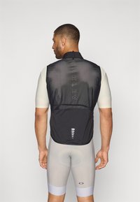 Veste légère noire avec col haut et accents réflecteurs, dotée d'un dos texturé, d'une poche zippée et d'un logo. Un short gris en dessous.