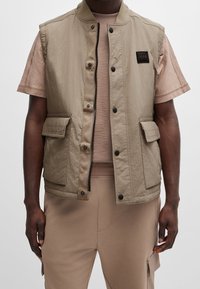 Chaleco sin mangas beige con una textura suave, cierre frontal, botones a presión y dos grandes bolsillos. Llevado sobre una camiseta ajustada de color marrón claro.