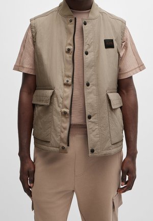 Beige mouwloos vest met een gladde textuur, voorrits, drukknoopjes en twee grote zakken. Gedragen over een lichtbruin getailleerd shirt.