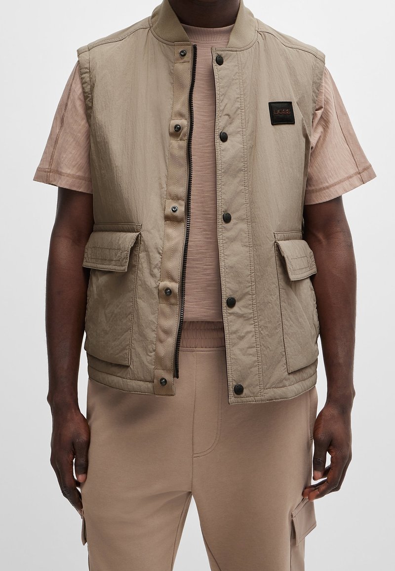 Chaleco sin mangas beige con una textura suave, cierre frontal, botones a presión y dos grandes bolsillos. Llevado sobre una camiseta ajustada de color marrón claro.