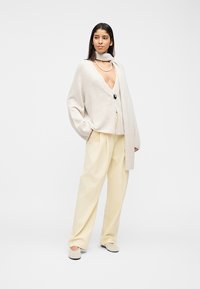 Γυναίκα που φοράει εκρού oversized ζακέτα με ένα κουμπί, μακρύ κασκόλ σε ταιριαστή απόχρωση, φαρδιά εκρού παντελόνια και επίπεδα παπούτσια, στέκεται με τα χέρια στις τσέπες.