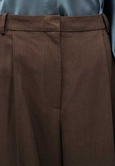Gros plan sur un pantalon marron taille haute avec passants pour ceinture, porté avec une chemise bleue rentrée.