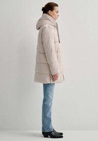 Giacca imbottita beige chiaro con cappuccio, caratterizzata da cuciture orizzontali e zip frontale. Abbinata a jeans azzurri chiari e stivaletti neri con tacco.