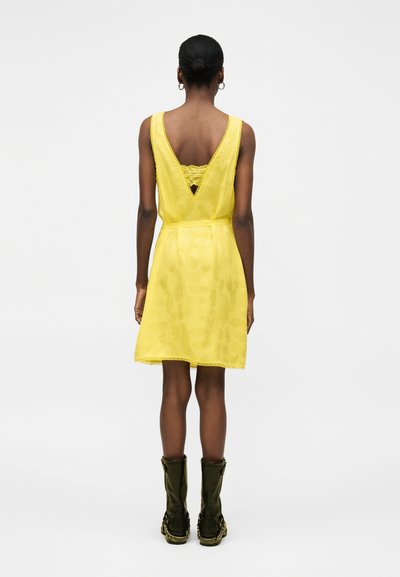 Γυναίκα με κίτρινο αμάνικο φόρεμα με V-πλάτη και δαντελένια λεπτομέρεια, σε συνδυασμό με σκούρα πράσινες μπότες μέχρι τη μέση της γάμπας, στέκεται μπροστά σε απλό φόντο.