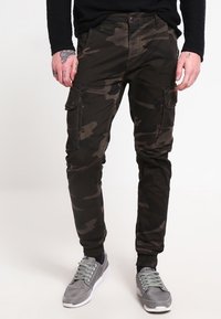 Osoba ubrana w ciemnozielone spodenki cargo w kamuflażu i szare sneakersy, stojąca na tle gładkiego jasnego tła.