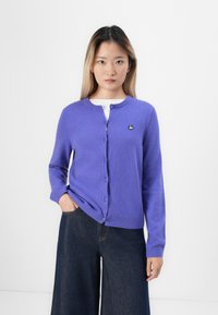 Jeune femme portant un cardigan violet à boutons et un pantalon large noir, se tenant avec une main dans sa poche devant un fond uni.