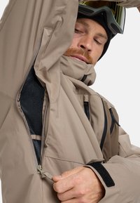 Beige waterproof jacket with mesh lining, black zippers, and Velcro cuffs. Model is adjusting a zipper while wearing ski goggles.