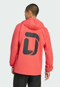 Roze jas met een capuchon, voorzien van een groot zwart logo op de achterkant. Het ontwerp heeft elastische manchetten en een gladde textuur.