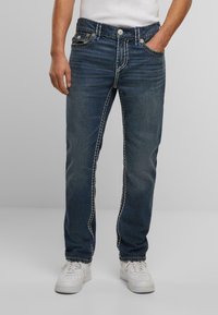 Vaqueros de denim azul con un corte recto, con costuras claras, cinco bolsillos y cierre de botón. Llevados con zapatillas blancas.