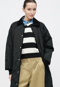 Czarny pikowany płaszcz z zatrzaskami i czarnym kołnierzem, noszony na czarno-białym paskowanym swetrze i jasnoniebieskiej koszuli z kołnierzykiem, zestawiony z khaki spodniami.